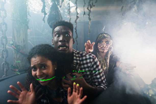 Young couple with zombie in halloween haunted house Two multi-ethnic young adults, an interracial couple, having fun in a halloween haunted house. They are on an amusement park ride, being chased by a demon or zombie in a dark, creepy tunnel. face paint halloween adult men stock pictures, royalty-free photos & images