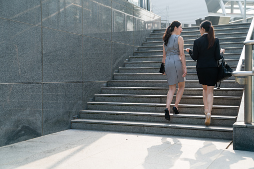 Two Asian office lady walk up the stairs and discuss with each other. On a back view. They both wearing high heels and formal suits dress carrying bags. Talking about the business stuff and costumers.
