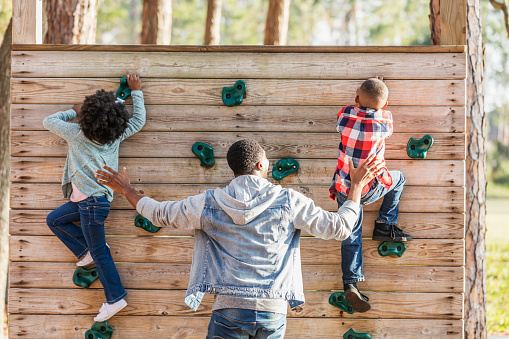 Rear view of an African-American father with his two young children playing at the park, on the playground. The 7 year old boy and 5 year old girl are on the climbing wall. Dad is behind them, ready to catch them if they fall.