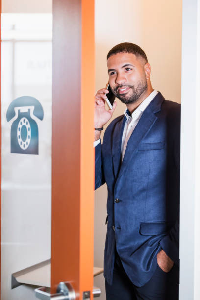 Businessman in booth talking on mobile phone A mid adult mixed race Hispanic and African-American man in his 30s with short hair, a beard and mustache, talking on his mobile phone, in a modern telephone booth to keep conversations private. pay phone on the phone latin american and hispanic ethnicity talking stock pictures, royalty-free photos & images