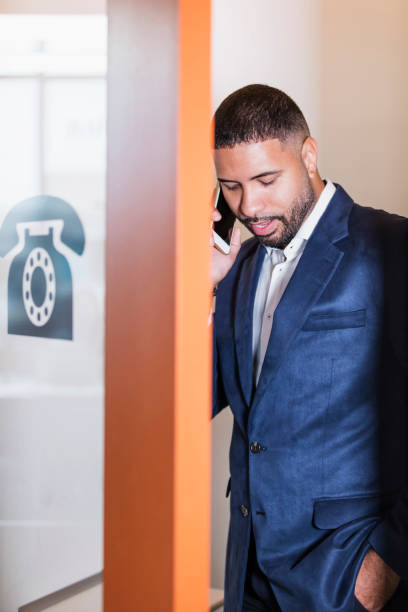 Businessman in booth talking on mobile phone A mid adult mixed race Hispanic and African-American man in his 30s with short hair, a beard and mustache, talking on his mobile phone, in a modern telephone booth to keep conversations private. pay phone on the phone latin american and hispanic ethnicity talking stock pictures, royalty-free photos & images
