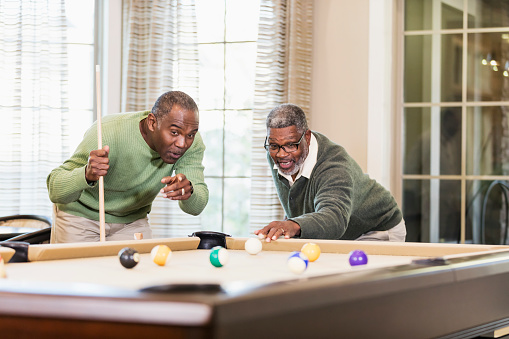 Two African-American men having fun shooting pool. The man about to his the cue ball with his cue stick is a senior man in his 60s. His friend, in his 50s, is pointing and talking, giving him advice.