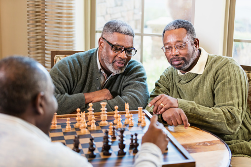 A group of three mature and senior African-American men in their 50s and 60s playing chess.  One man is moving a piece while his friends watch and discuss strategy.