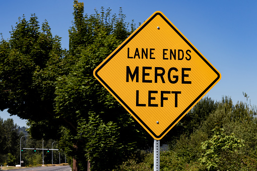 Lane ends merge left sign with trees and a blue sky