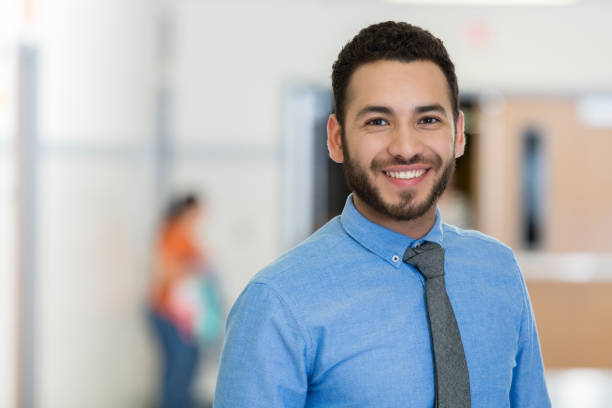 Young male educator stands proudly in school building A smiling young man wears a tie as he stands in the hallway of his school building and smiles proudly for the camera.  He is a school staff member. necktie photos stock pictures, royalty-free photos & images
