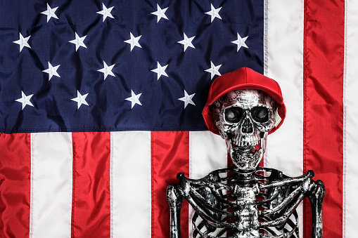 An empty humorous hillbilly skeleton voter wearing a symbolic red hat standing in front of an American flag background.