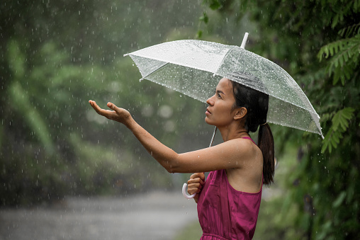 Beautiful young woman so sad as she holds out her palm to catch falling water. She is holding an umbrella over her head. Horizontal shot. Rainy day asian woman wearing a pink coat outdoors.