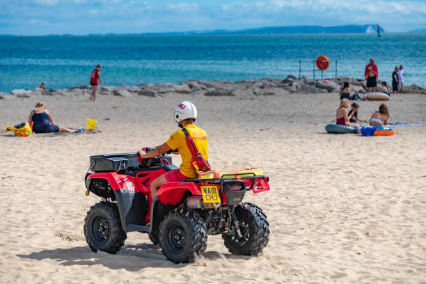 RNLI lifeguard on a quadbike on the beach Poole, UK - August 21 2018: A male RNLI lifeguard rides on a quadbike patrolling the beach at Sandbanks in Poole, Dorset. Other people on the beach sunbathing. sandbanks poole harbour stock pictures, royalty-free photos & images