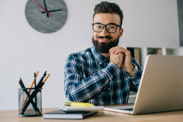 istock handsome smiling businessman in eyeglasses looking at camera while sitting at workplace with laptop 1023378774