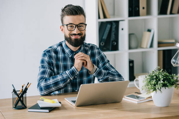 istock bearded smiling businessman in eyeglasses looking at camera while sitting at workplace with laptop 1023378800