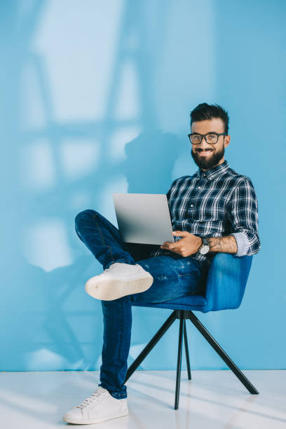 istock smiling man using laptop and sitting in armchair, on blue 1023378936
