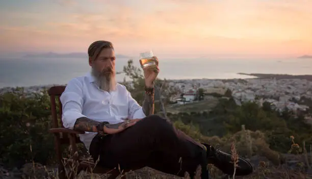 Attractive man with a beard is relaxing with a glas of whiskey in his hand in a beautiful landscape