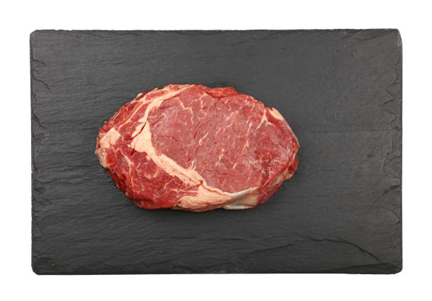 Close up one raw ribeye beef steak on black slate board isolated on white background, elevated top view, directly above