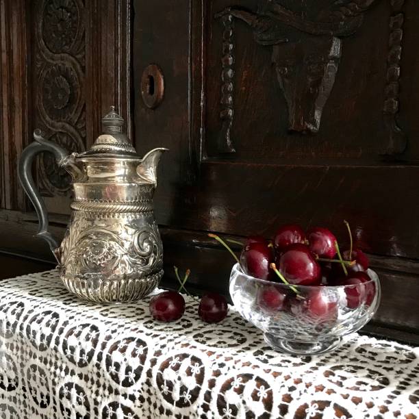 A still life with a silver water jug and red cherries on a court cupboard A small silver chased water jug with a black ebony handle and dark red cherries in a glass bowl on a 17th century oak court cupboard antique fruit bowl stock pictures, royalty-free photos & images