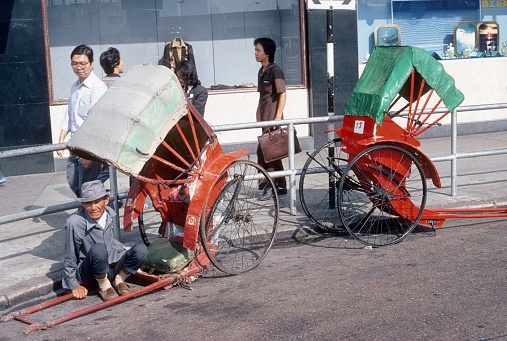 Hong Kong, China, 1980. An old rickshaw driver is waiting by the roadside for customers. Furthermore: passers-by, sidewalk and buildings.