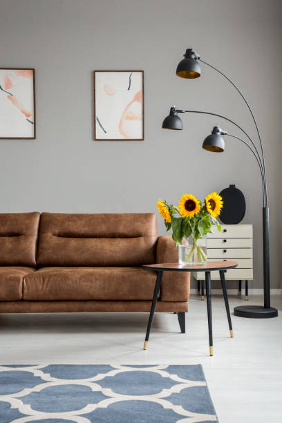 istock Lamp and table with sunflowers next to leather couch in grey flat interior with posters. Real photo 1023481044