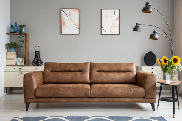 кожаный диван рядом со столом с подсолнухами в сером интерьере гостиной с плакатами. реальное фото - sofa стоковые фото и изображения
