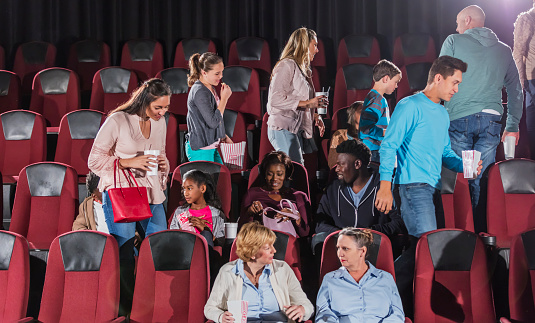 A large, multi-ethnic group of people getting up from their seats in a movie theater, after watching a movie. They are mixed ages, from children to mature adults, including families and friends.