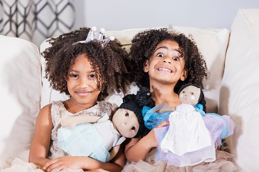 Two cute mixed race African-American and Hispanic girls, sisters 3 and 5 years old, playing together, sitting side by side on a sofa, holding dolls in their laps, wearing princess dresses and tiaras, smiling at the camera.