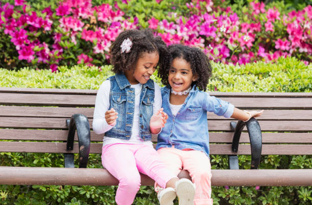 Two cute mixed race African-American and Hispanic girls, sisters 3 and 5 years old, sitting together side by side on a park bench, laughing and playing.