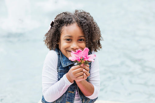 A beautiful mixed race African-American and Hispanic girl, 5 years old, holding pink flowers, smiling at the camera.