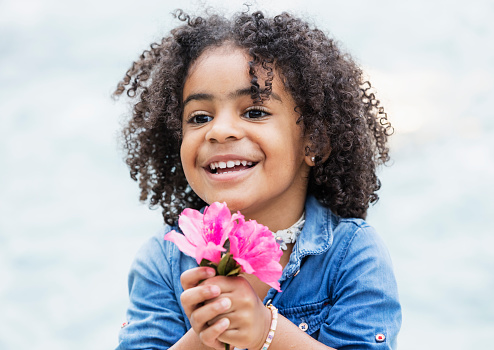 A beautiful mixed race African-American and Hispanic girl, 3 years old, holding a pink flower, smiling.
