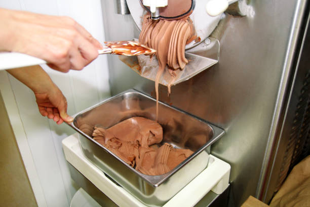 istock Confectioner in chef uniform is working on ice cream maker machine. Producing black chocolate ice cream flavors and it falls into steel container. Industrial preparation of italian creamy ice cream. 1023505948