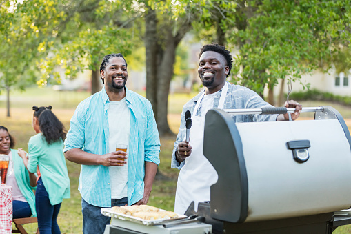 Two African-American men in their 30s, brothers, enjoying a cookout in their back yard. They are standing at the grill, one of them wearing an apron and holding tongs, and the other holding a drink. They are smiling and looking at the camera.