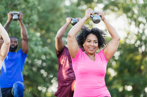 A multi-ethnic group of people of mixed ages in an exercise class outdoors at the park, lifting kettlebells above their heads. The focus is on a senior African-American woman in her 60s.