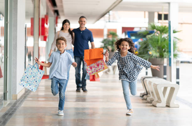 Beautiful siblings having fun at the mall running and holding shopping bags Beautiful siblings having fun at the mall running and holding shopping bags and parents walking behind them embracing each other shopping bag photos stock pictures, royalty-free photos & images