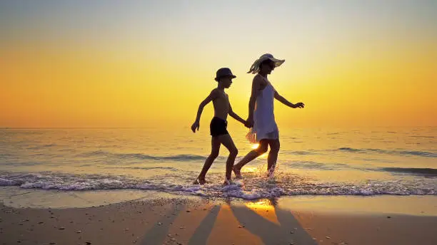 Photo of Family on summer vacation. Mother in white dress and son wearing hat walk on an epmty beach at vibrant sunset