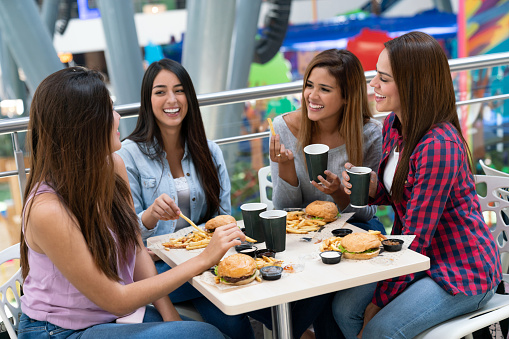 Latin american group of female friends having fun at the mall eating hamburgers in the food court while talking and laughing