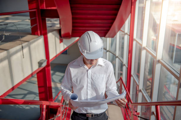 istock Young Asian male engineer or architect holding blueprints wearing protective safety helmet on red stair at construction site. Civil engineering, Architecture and building construction concept 1023560062