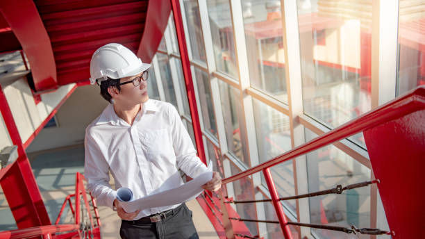 istock Young Asian male engineer or architect holding blueprints wearing protective safety helmet on red stair at construction site. Civil engineering, Architecture and building construction concept 1023560120