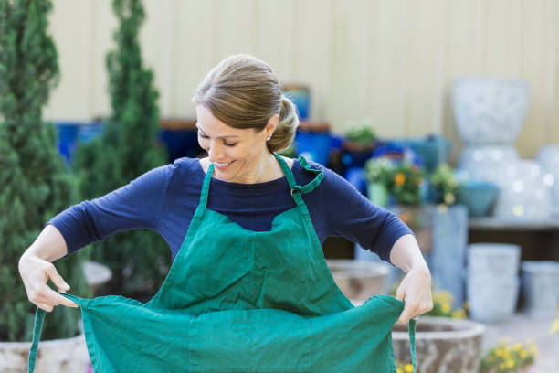 A mature woman in her 40s working in a plant nursery or garden center. She is smiling as she puts on a green apron.