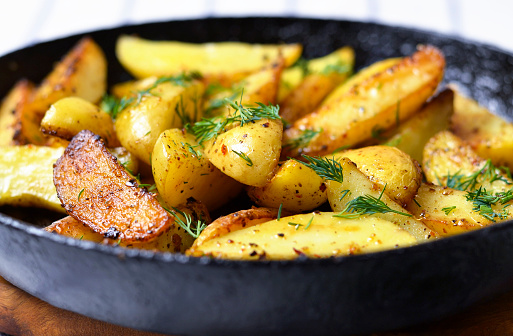 Closeup of fried potato wedges with dill, garlic and pepper, decorated in a rustic style. Rustic fried potatoes in a cast iron skillet at home.