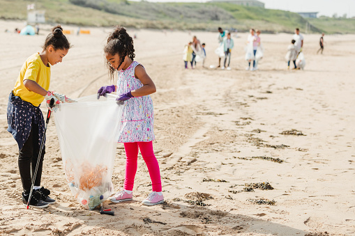 Young girls collecting rubbish off a beach. Plastic containers, bottles in their bag. They are wearing protective gloves and using a mechanical grabber.