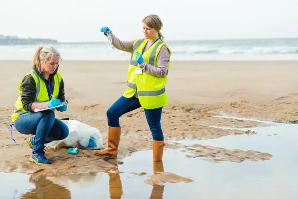 Two females working on the beach by a pool of water collecting and analysing samples. They are wearing protective gloves and reflective clothing