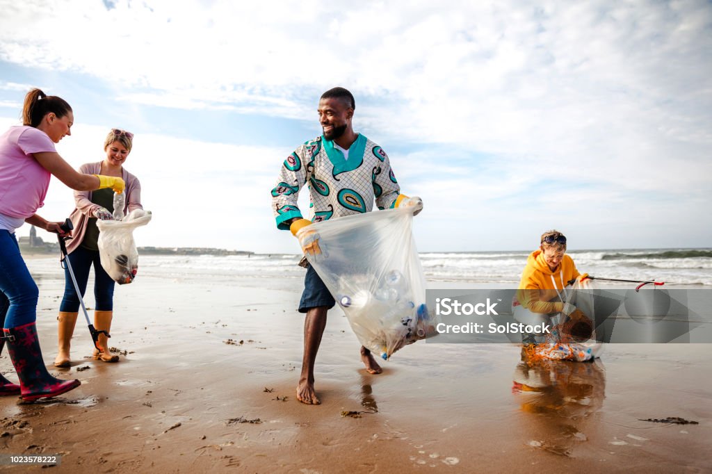 Activists Working Together Making a Difference Collecting rubbish off a beach. Plastic containers, bottles in their bag. They are using a mechanical grabber. Three women and a man working together Cleaning Stock Photo