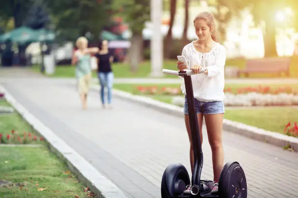 A young beautiful girl with a smartphone is riding a segway in a park. Healthy lifestyle.