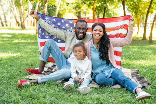 アフリカ系アメリカ人の両親と娘は、公園でのピクニックでアメリカの国旗を保持している笑みを浮かべてください。 - preschooler child offspring purity ストックフォトと画像