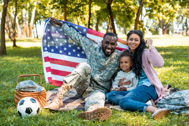 felice soldato afroamericano in uniforme militare e famiglia che tiene bandiera americana nel parco - family american culture african culture black foto e immagini stock