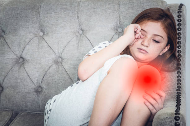 istock Young daughter sitting on sofa with knee pain. Red highlight at knee. 1023588524