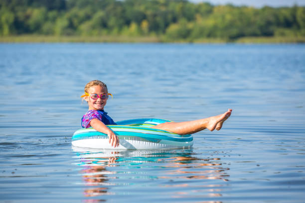 Side view of a young girl laying on her back in an inner tube float toy, floating on the water at the lake on a summer day. The girl is wearing a rash guard swim shirt and swim goggles, and she is smiling at the camera.