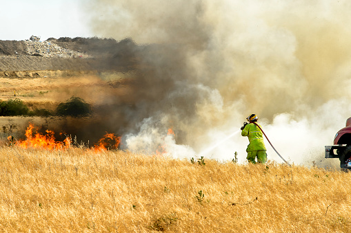 Smoke and flames from dry grass wildfire burning near rural farm, with unrecognizable firefighter hosing down the fire.