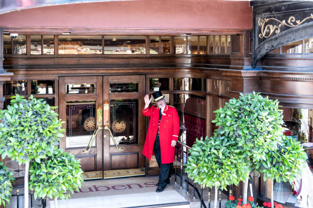 Porter, doorman in traditional attire, clothing, hat, red gown, coat standing at Rubens hotel, inn entrance waving hand London, UK - June 22, 2018: Porter, doorman in traditional attire, clothing, hat, red gown, coat standing at Rubens hotel, inn entrance waving hand door attendant photos stock pictures, royalty-free photos & images