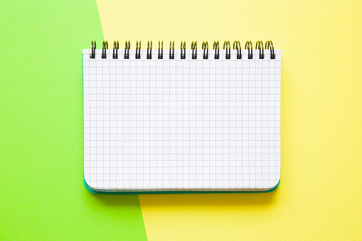 White squared notebook on the green, yellow work table. Empty place for daily plans, important information, ideas, memories or other text on the grid paper. Bright colors. Top view. Flat lay.