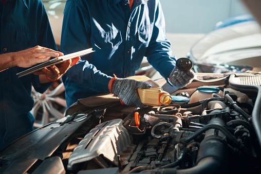 Mechanic pouring oil in car engine when his colleague using tablet computer