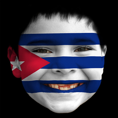 A lovely cheerful six year old boy smiling with broken incisors and half grown incisor teeth with Cuba flag painted on his face. Alternating stripes of blue and white and a red equilateral triangle at the hoist, within which is a white five-pointed star.