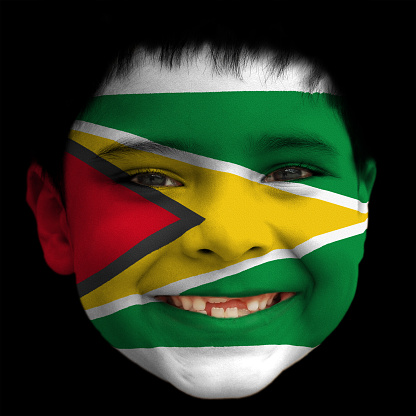 A lovely cheerful six year old boy smiling with broken incisors and half grown incisor teeth with Guyana flag painted on his face. Red, Yellow, Green, White and Black colours. Geometric shapes, Triangles.